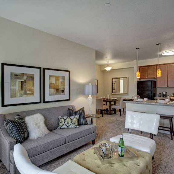 A living room with a gray couch, white chairs, a yellow footrest, wall art, and view of the kitchen and dining room at Solara Apartment Homes. 