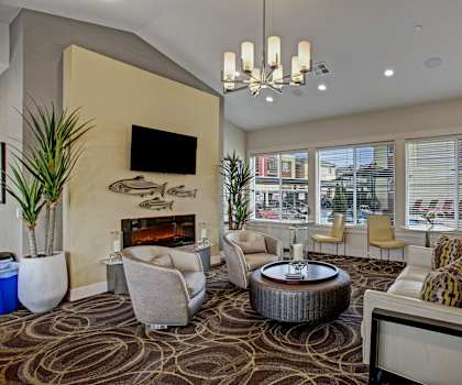 The resident lounge with tan leather couches, plush chairs, and tables and chairs by the large windows, and a cozy fireplace with a TV and fish decorations above and two plants on either side at Riverside 9 Apartment Homes. 