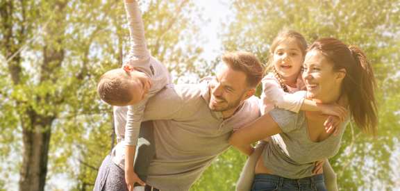 family of four piggyback with kids in park with trees