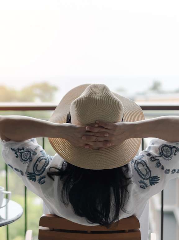 A woman in a hat is sitting on a chair with her back to the camera, looking out of a balcony window.