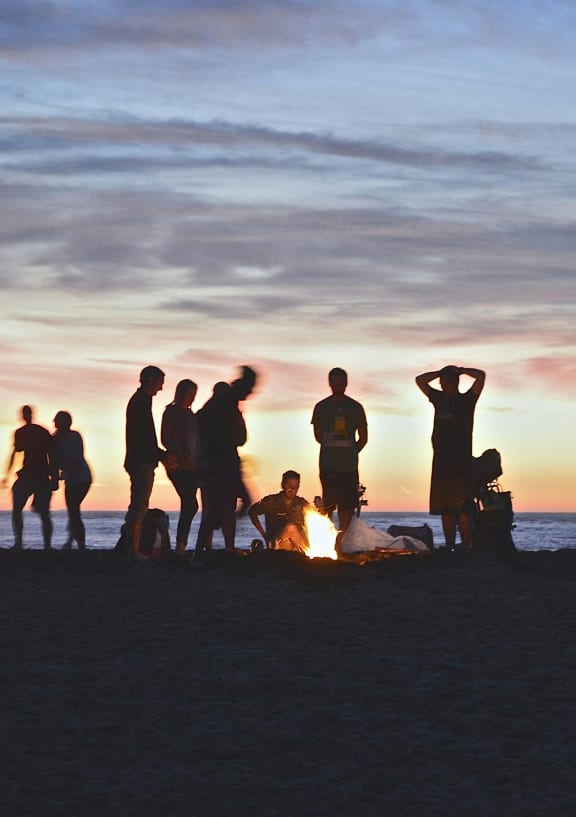 a group of people standing around a fire on the beach at sunset