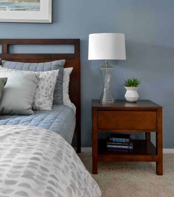 a bedroom with a bed and a nightstand with a lamp and a plant at The Villas at Main Street, Ann Arbor, MI