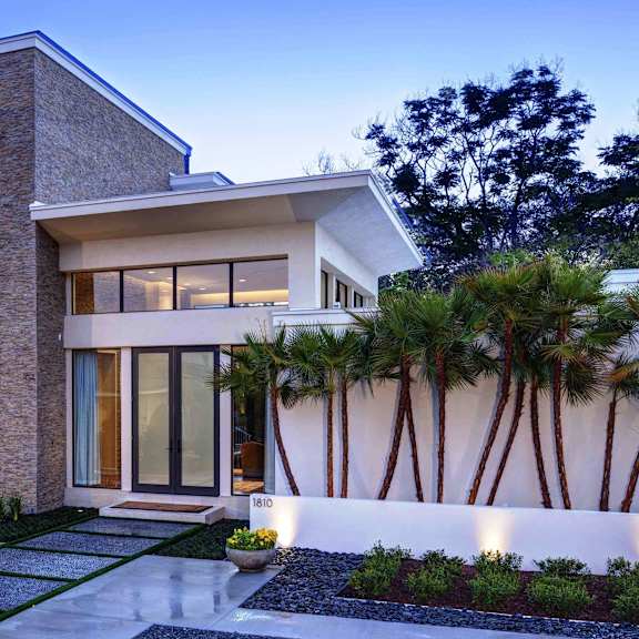 A modern house with a stone facade and a white wall with palm trees in front.