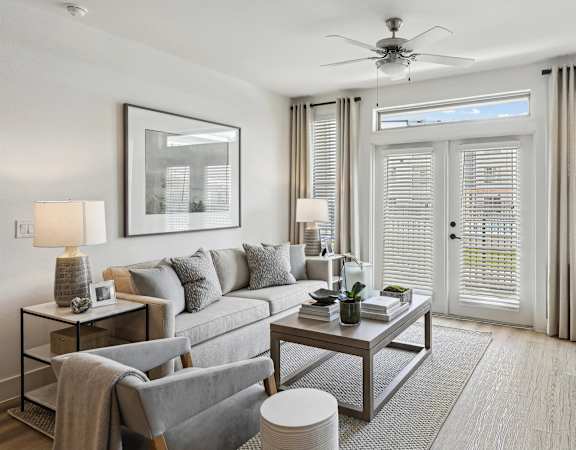 A living room with a grey sofa, a grey armchair, a glass table with a vase and a book on it, and a ceiling fan.