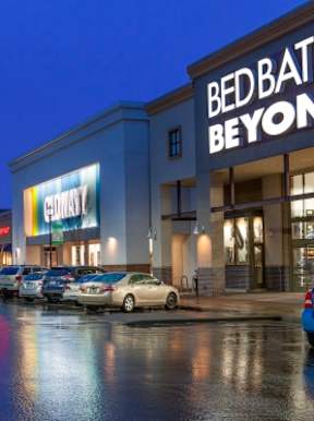 Bed Bath and Beyond storefront at night with cars parked in front.