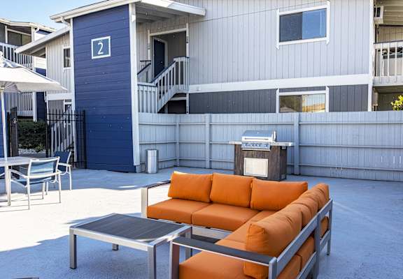 Outdoor lounge with umbrella-covered tables, gas BBQ, and an oversized orange patio couch and coffee table.at The Lakes Apartments, Moses Lake, WA
