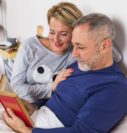 an older man reading a book to a woman in bed