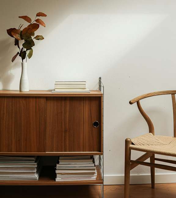 A wooden chair with a beige cushion sits next to a wooden cabinet with a vase of leaves and books on top.