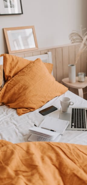 a laptop on a bed with orange pillows and a cup of coffee