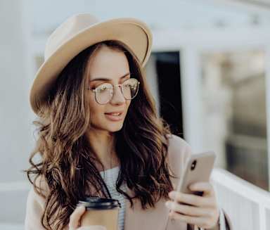 A woman in a beige coat and hat is holding a coffee cup and looking at her phone.