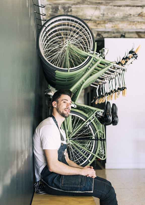 A man sits in front of a wall with a green bicycle wheel mounted on it.