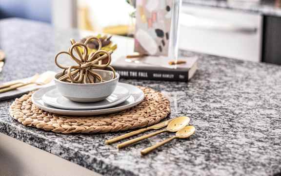 A bowl of noodles sits on a plate with chopsticks on a granite countertop.