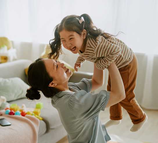 A woman is lifting a girl in the air in a living room.