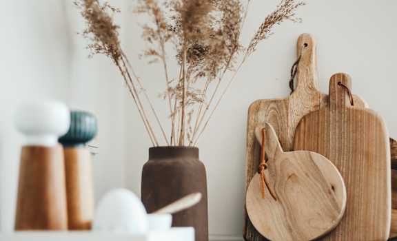 A wooden cutting board and a vase with dried plants are on a reflective surface.