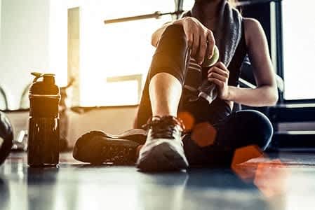 A person is sitting on the floor with a water bottle and a towel, wearing black workout clothes and sneakers.
