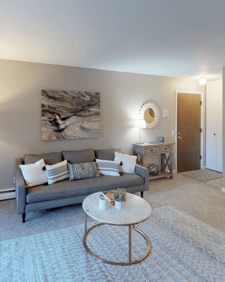 A grey couch with white pillows sits in a living room with a round mirror and a painting on the wall.