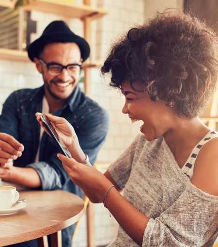 A man and a woman are sitting at a table, with the woman holding a smartphone and both smiling.