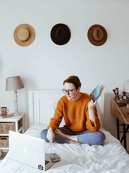 a woman sitting on her bed reading a book and using her laptop