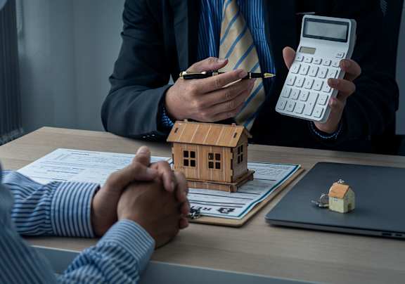A person in a suit is holding a calculator and appears to be discussing a financial matter with another individual.