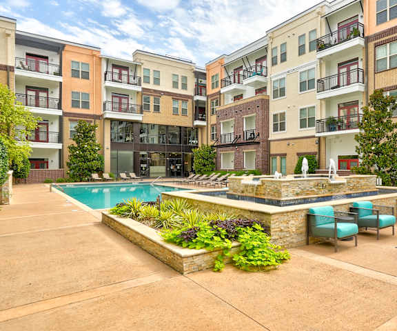 The large, turquoise outdoor pool with a fountain, pool chairs, tables, and turquoise chairs with the apartments in the background at the Edge at Midtown Apartment Homes. 