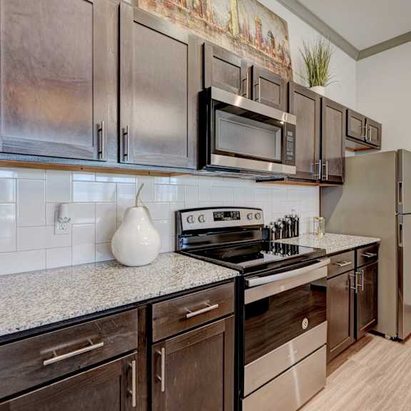 A large, modern kitchen with stainless steel appliances, with a ceramic pear, kitchen utensils, marble countertops with lots of room, and large, dark wooden cabinets at the Encore Apartments. 