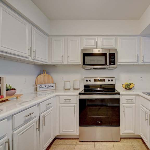 A large, modern kitchen with stainless-steel appliances, white marble countertops with lots of room, and large white wooden cabinets with plants and decorations including a cookbook at Villas at Preston Lakes Apartment Homes.