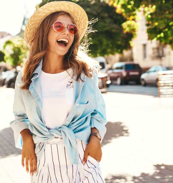 A woman in a straw hat and striped pants is laughing on a sunny street.