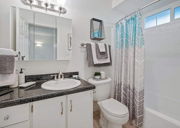 One-Bedroom Apartments in Hayward, CA - Glen Oaks - Bathroom with Gray Granite Countertop Sink, White Cabinetry, Above Mirror Cabinet with Overhead Lighting, and a Shower with a Tub and a Blue/Gray Shower Curtain.
