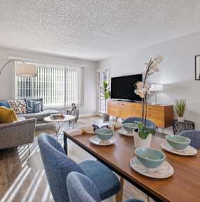 Hayward, CA, Apartments - Glen Oaks - Dining Area with Table and Plush Chairs Connected to Living Room with Gray Couch, Small Round Coffee Table, TV Table with Cabinets, Wood-Inspired Flooring, and a Large Window with White Blinds.