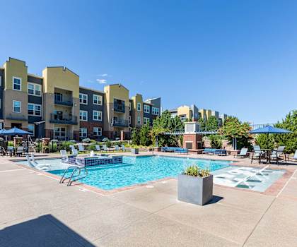 A swimming pool surrounded by chairs and umbrellas in front of apartment buildings.