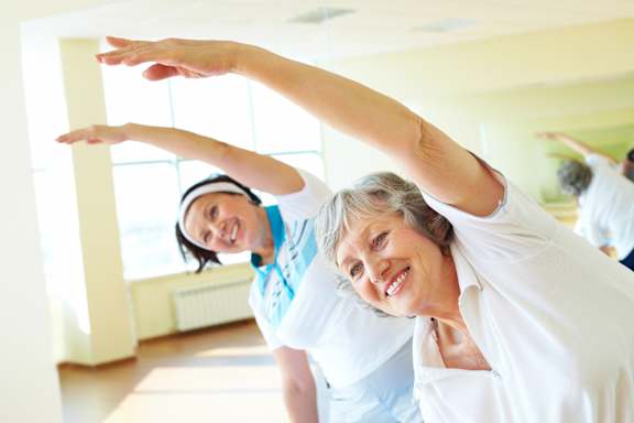 A woman in a white shirt is stretching her arms upwards in a bright room.