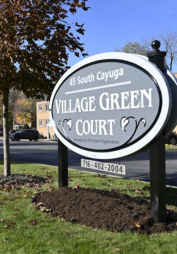 A sign for Village Green Court stands in front of a tree and a building.