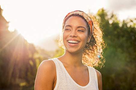 A woman with curly hair and a white tank top is smiling in a sunny outdoor setting.