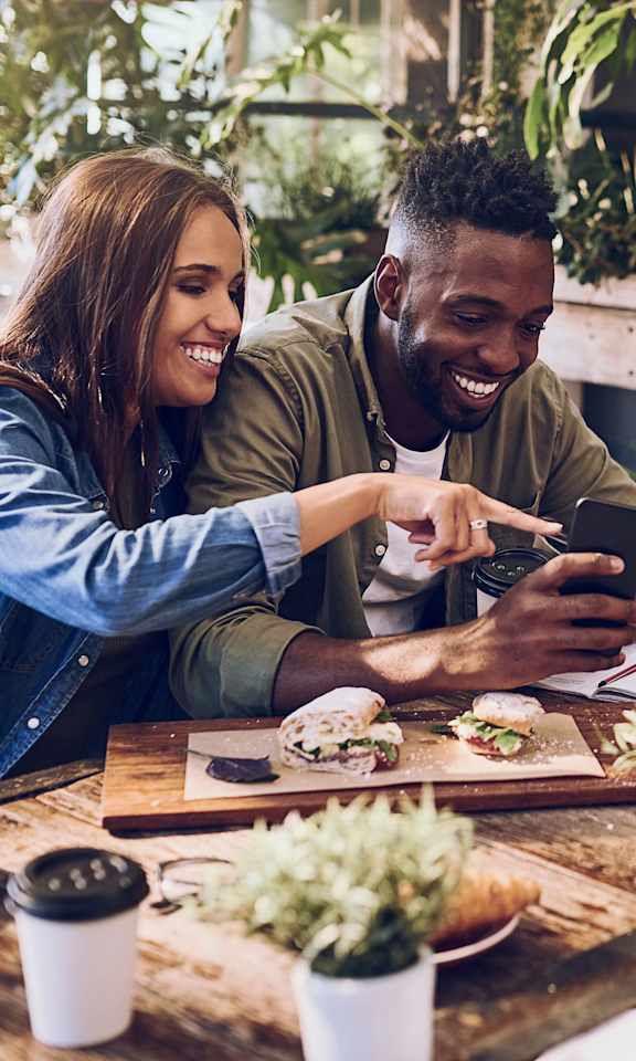 Three people are sitting at a table with food and drinks, looking at a smartphone.