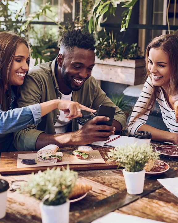 Three people are sitting at a table with food and drinks, looking at a smartphone.