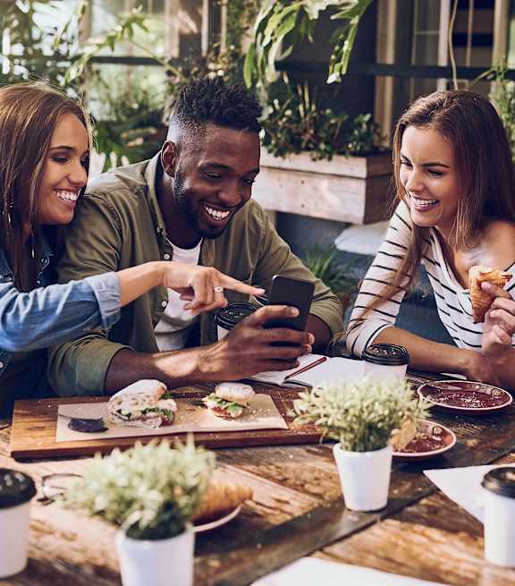 Three people are sitting at a table with food and drinks, looking at a smartphone.