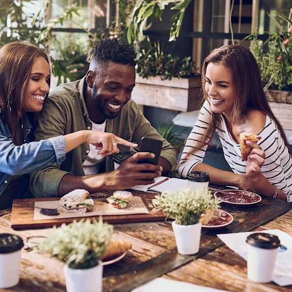 Three people are sitting at a table with food and drinks, looking at a smartphone.