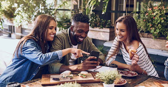 Three people are sitting at a table with food and drinks, looking at a smartphone.