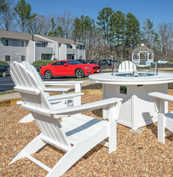 A white table and chairs set is set up on a gravel area with a red car and houses in the background.