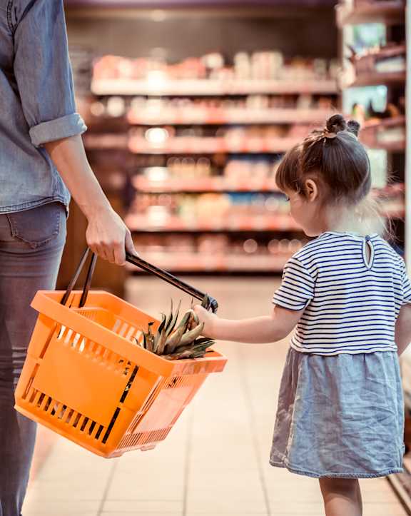 A woman and a little girl are shopping, the girl is holding a basket full of groceries.