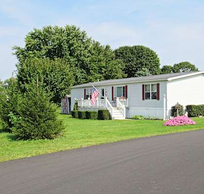 A white house with a flag on the front porch is surrounded by greenery and a paved road.