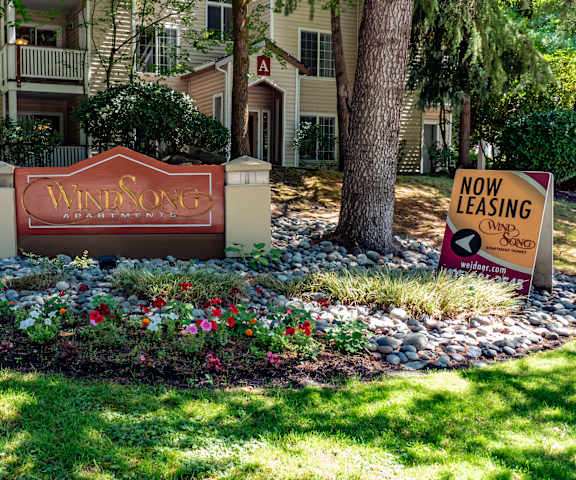 The exterior sign with trees and surrounded by greenery with the buildings in the background of Windsong Apartment Homes. 