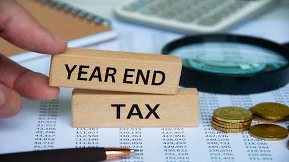 A hand holds a block with the words "YEAR END TAX" in front of a calculator and some coins.