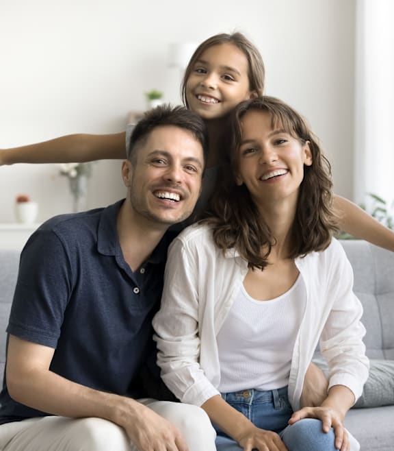 A family of three is sitting on a grey couch in a bright room.