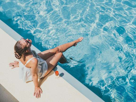 A woman is sitting by the poolside with her legs in the water.
