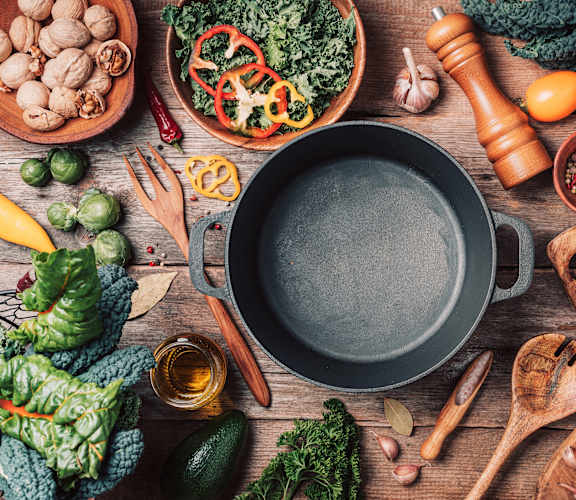A variety of fresh vegetables and herbs are arranged around a large pot on a wooden surface.