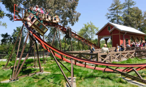 People riding a wooden roller coaster on a sunny day.