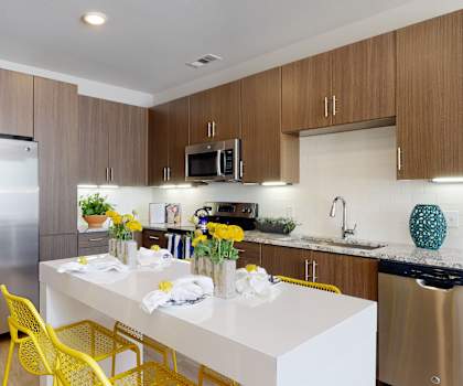 A large, modern kitchen with stainless steel appliances, kitchen utensils, a turquoise vase, white marble countertops with lots of room, a kitchen island with barstools and four places set for a meal with two vases full of yellow flowers, and large, medium wooden cabinets at the Flats at San Tan Apartment Homes. 
