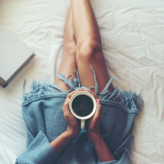 A person is lying on a bed with their legs up, holding a cup of coffee and a book.