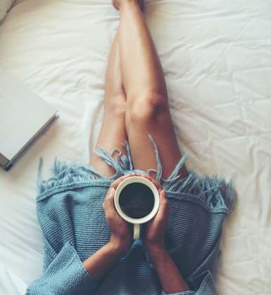 A person is lying on a bed with their legs up, holding a cup of coffee and a book.